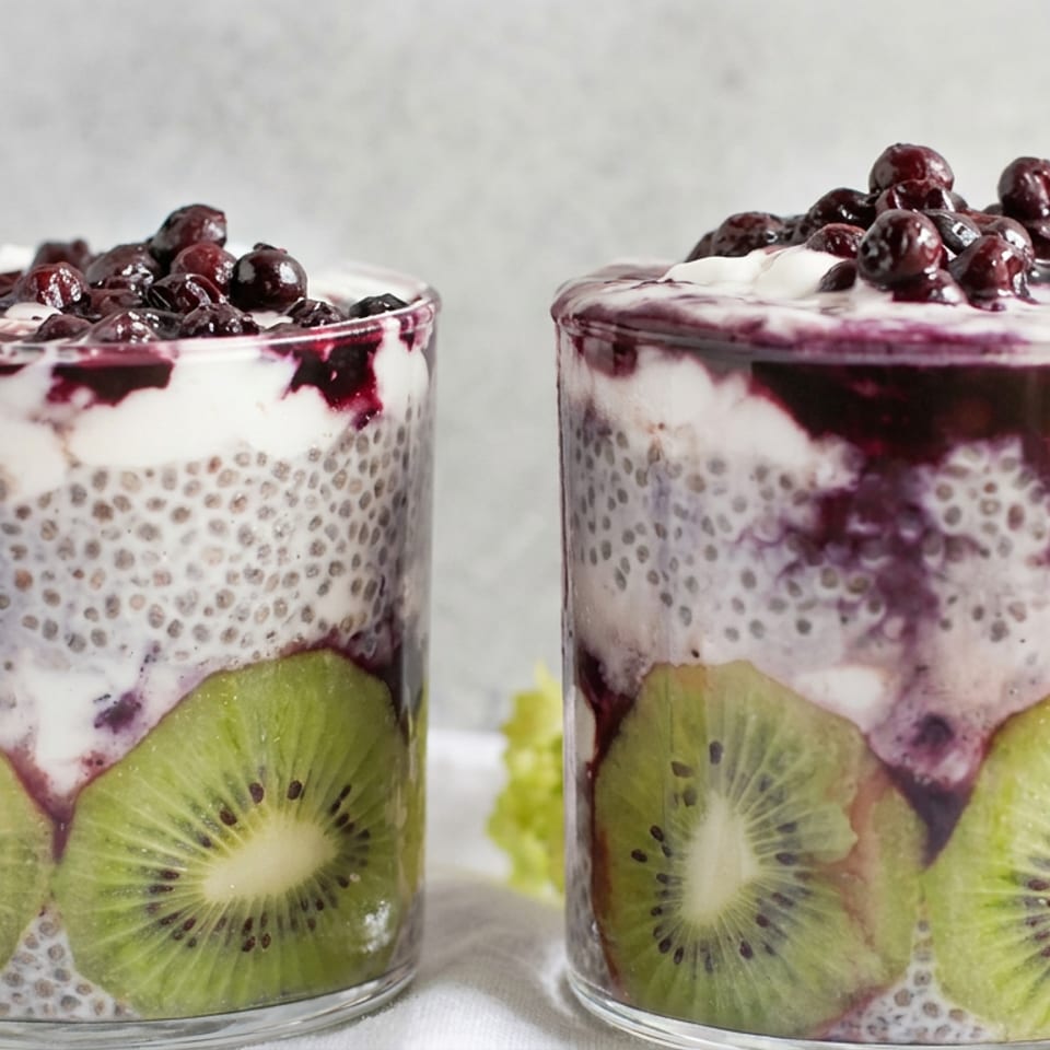 Two glasses of layered chia pudding with kiwi slices, yogurt, and blueberry compote topped with blueberries.