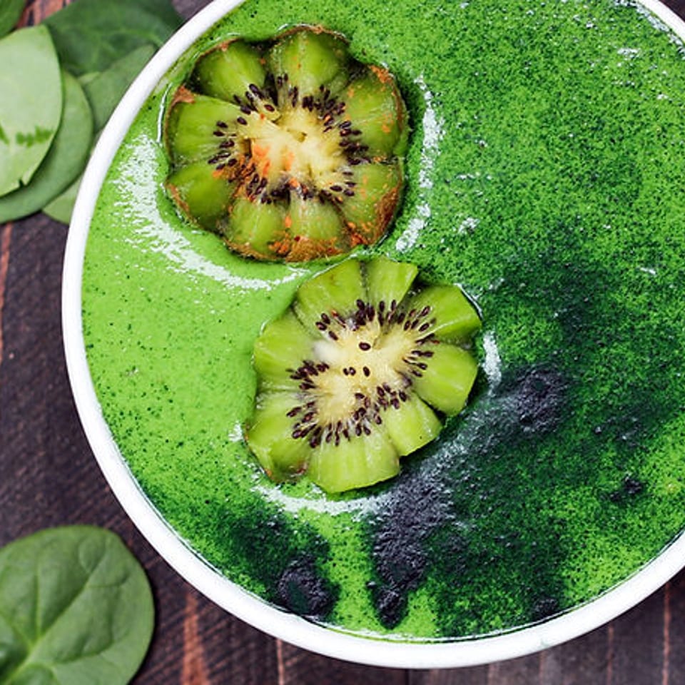 Green smoothie bowl topped with kiwi slices, leafy greens, and dark berries on a rustic wooden table.